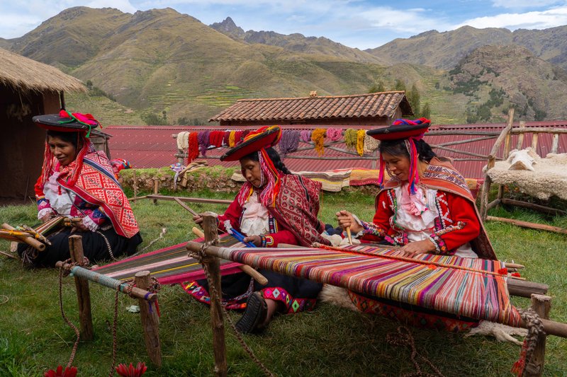 Three local female weavers in colourful traditional local dress including festooned hats, weaving colourful alpaca wool on the ground, Chumbe Community